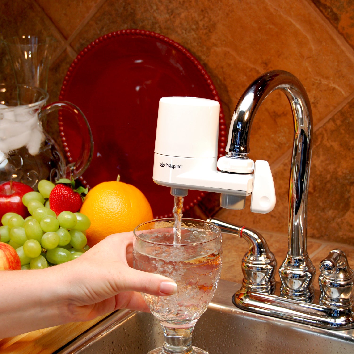 Hand pouring water from a faucet-mounted water filter into a glass, with fruits on a kitchen counter.