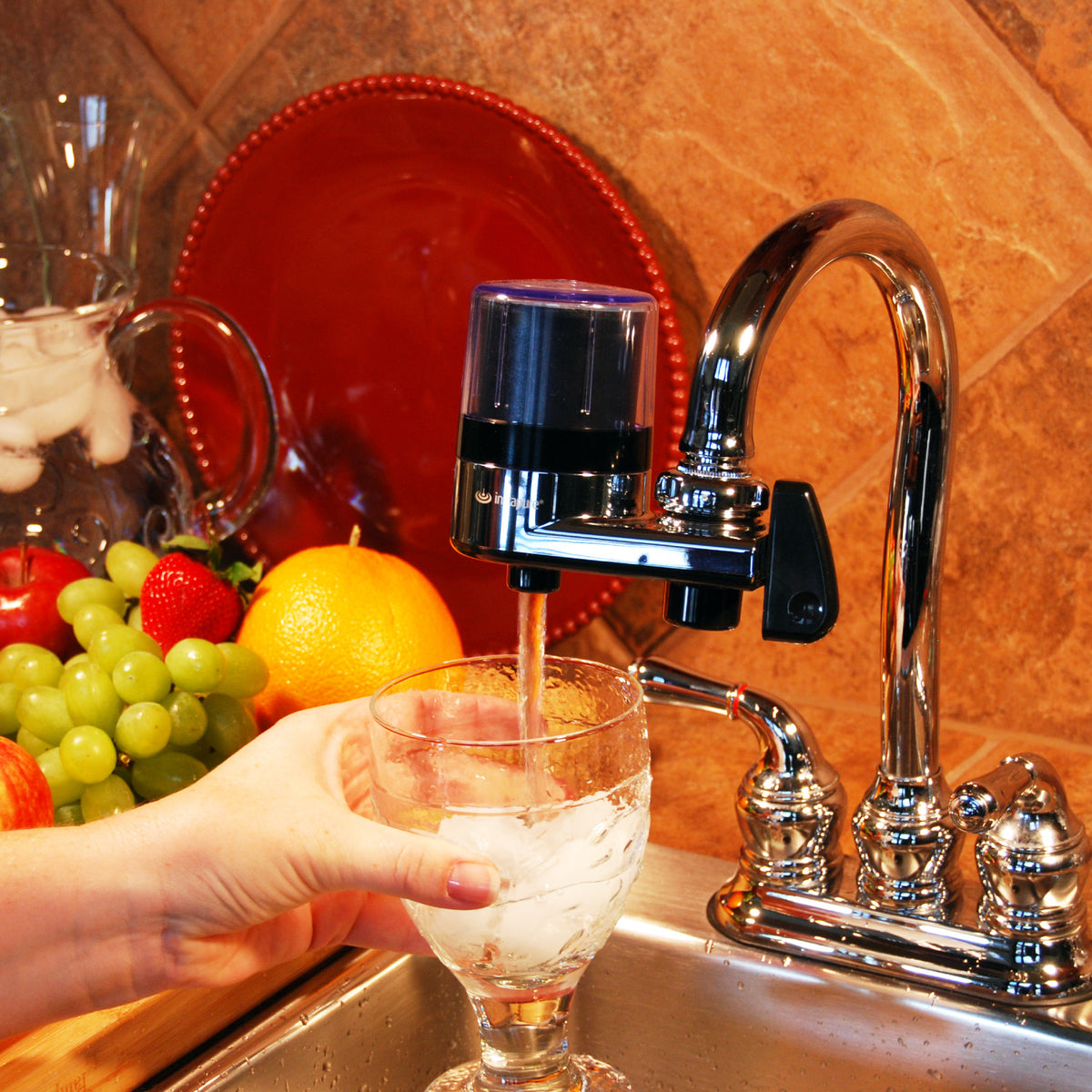 Person pouring water from a F2 faucet-mounted water filter into a glass, with fruits on a counter.