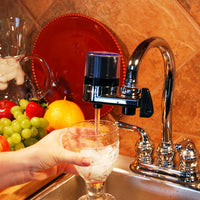 Person pouring water from a F2 faucet-mounted water filter into a glass, with fruits on a counter.