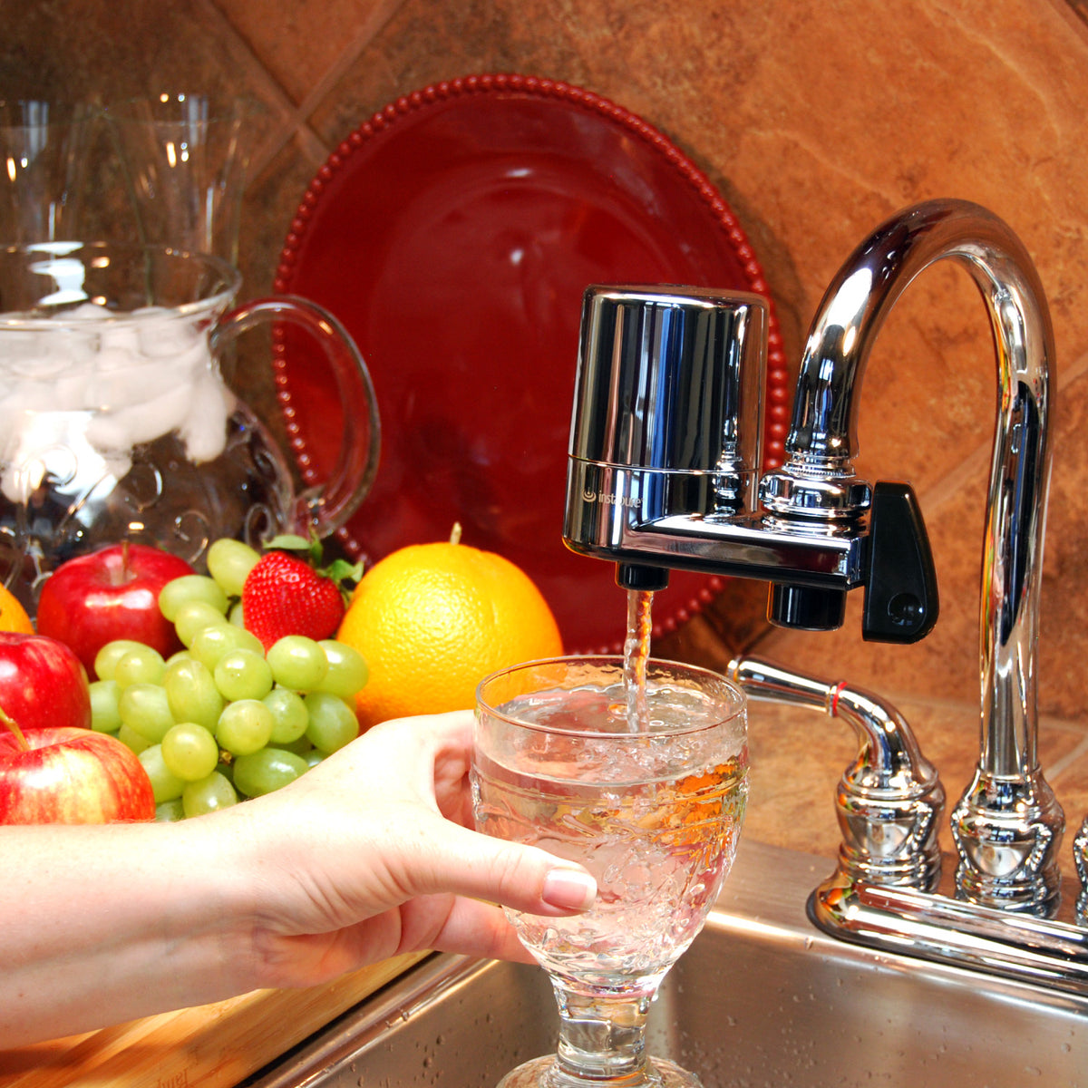 Water being poured from a F2 faucet into a glass with fruits and a red plate in the background.