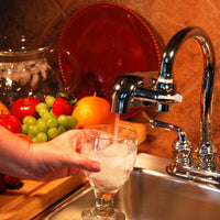 Person filling a glass with water from a F8 Chrome System surrounded by fruits on a kitchen counter.